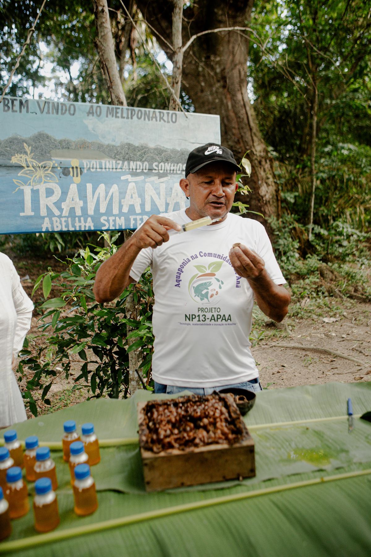 Homem apresentando produtos regionais a visitantes, ao lado de uma placa de comunidade