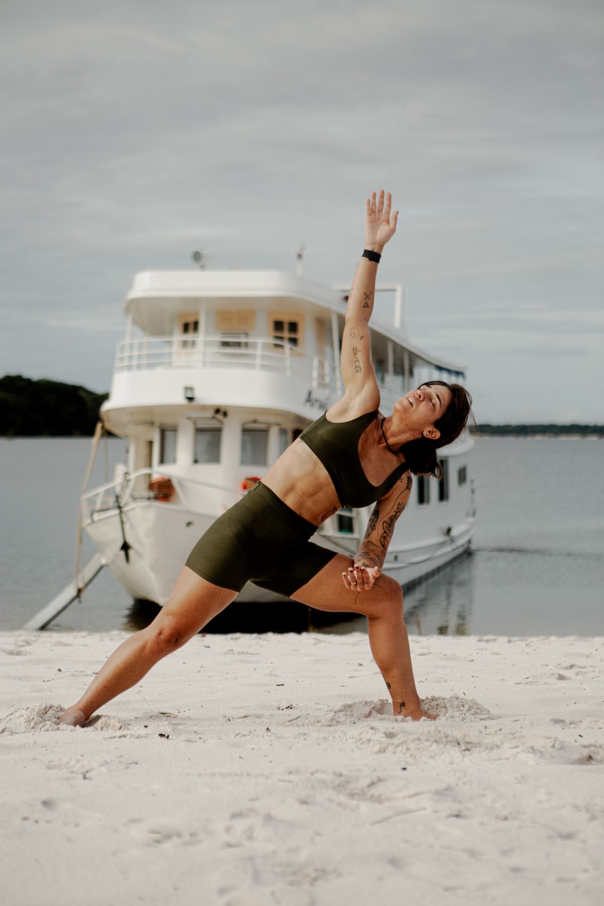 Mulher numa postura de yoga na praia, com um barco ao fundo